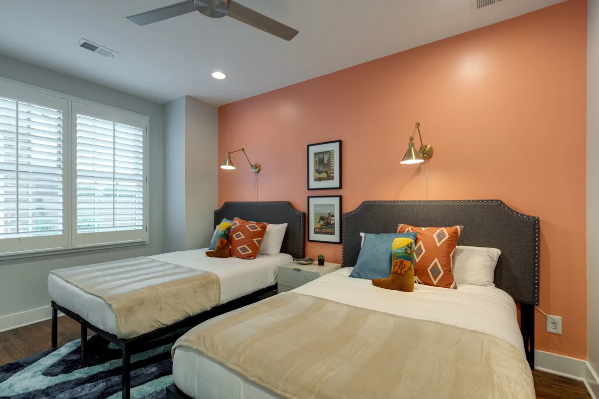 A cheerful bedroom with two beds adorned with decorative pillows, colorful wall art, stylish reading lamps, and large windows with shutters allowing natural light to filter in. The layout feels ideal for friends or families sharing the space.