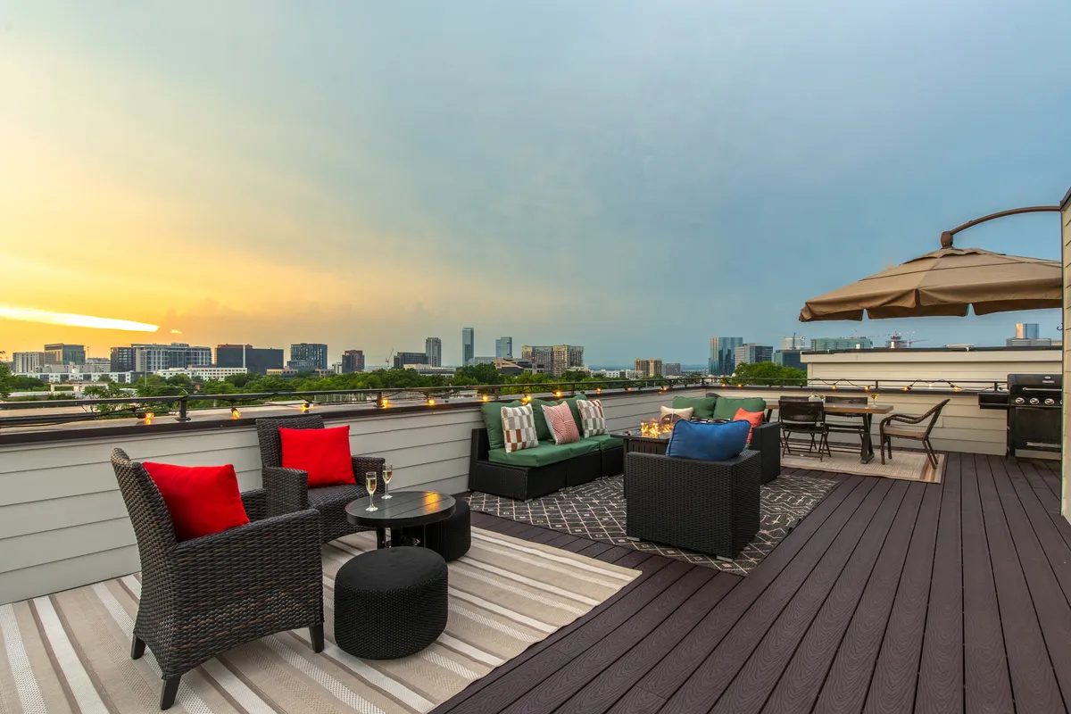 The image features a stylish outdoor rooftop seating area adorned with colorful decorative cushions and an inviting dining setup, all overlooking the Nashville skyline at sunset. The scene radiates warmth and comfort, perfect for gathering with friends to relax, dine al fresco, or take in the vibrant city views as day turns to night.