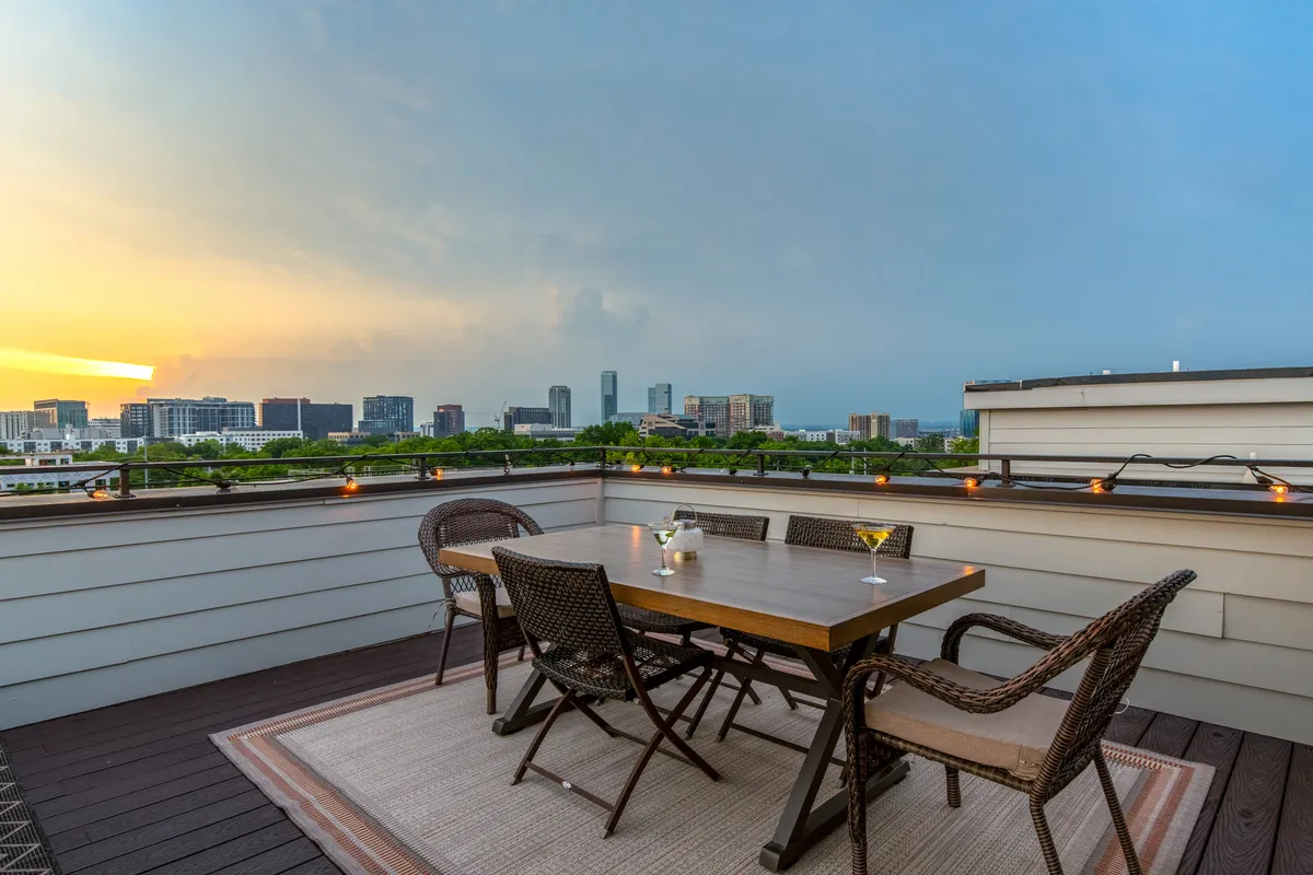 A dining table is set beneath string lights on a rooftop, with the sun setting behind the Nashville city skyline. The scene is intimate and inviting, ideal for outdoor dining and evening drinks as twilight descends.