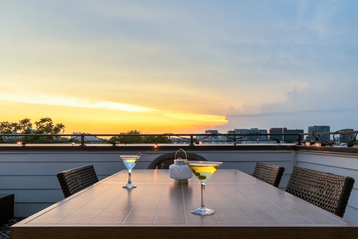 Cocktail glasses sit atop a dining table on a rooftop, with the sun dipping below the city skyline. The mood is sociable and tranquil, suggesting lively gatherings as day turns to night.