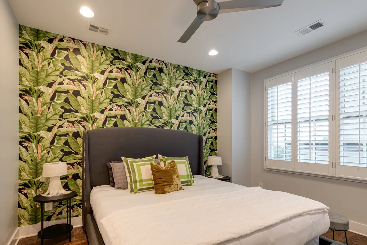 A stylish bedroom featuring bold tropical wallpaper, a comfortable bed with decorative pillows, and nightstands with modern lamps. The space feels tranquil and inviting, perfect for relaxation and privacy. Natural light filters in from the windows, enhancing the peaceful atmosphere.