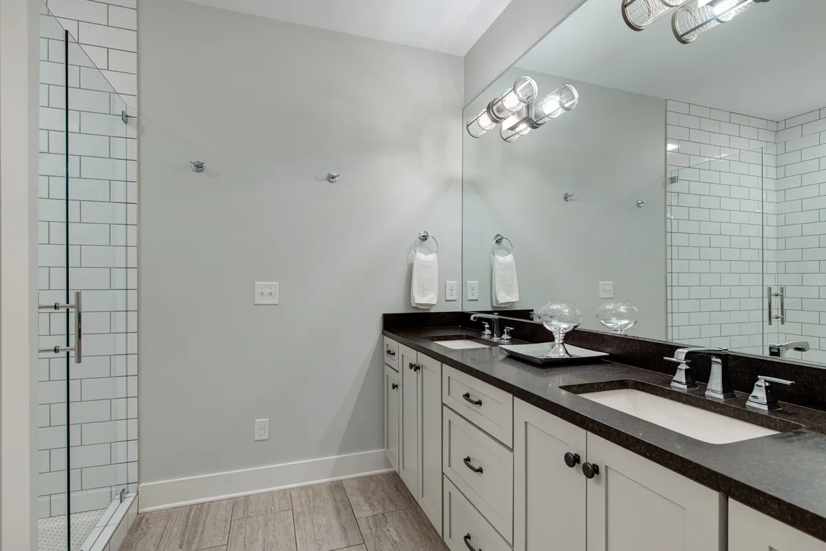 A modern bathroom with a double sink vanity, large mirror, and a glass-enclosed shower. The fresh, inviting ambiance is complemented by ample space and morning light, emphasizing relaxation and privacy.