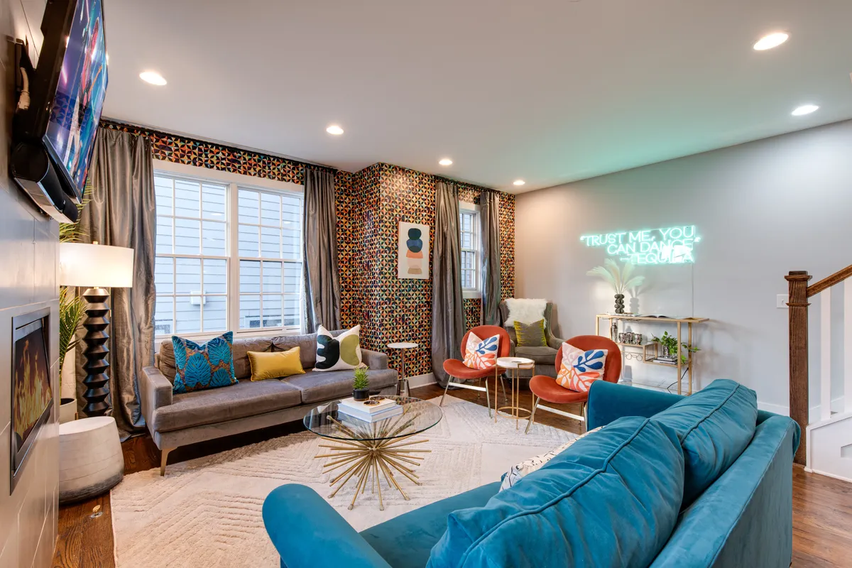 A lively second-floor living area with a colorful accent wall, a plush sofa adorned with decorative cushions, a fun neon sign, and stylish chairs arranged for conversation. The room’s ambiance is playful and energetic, ideal for group gatherings, celebrations, or movie nights.