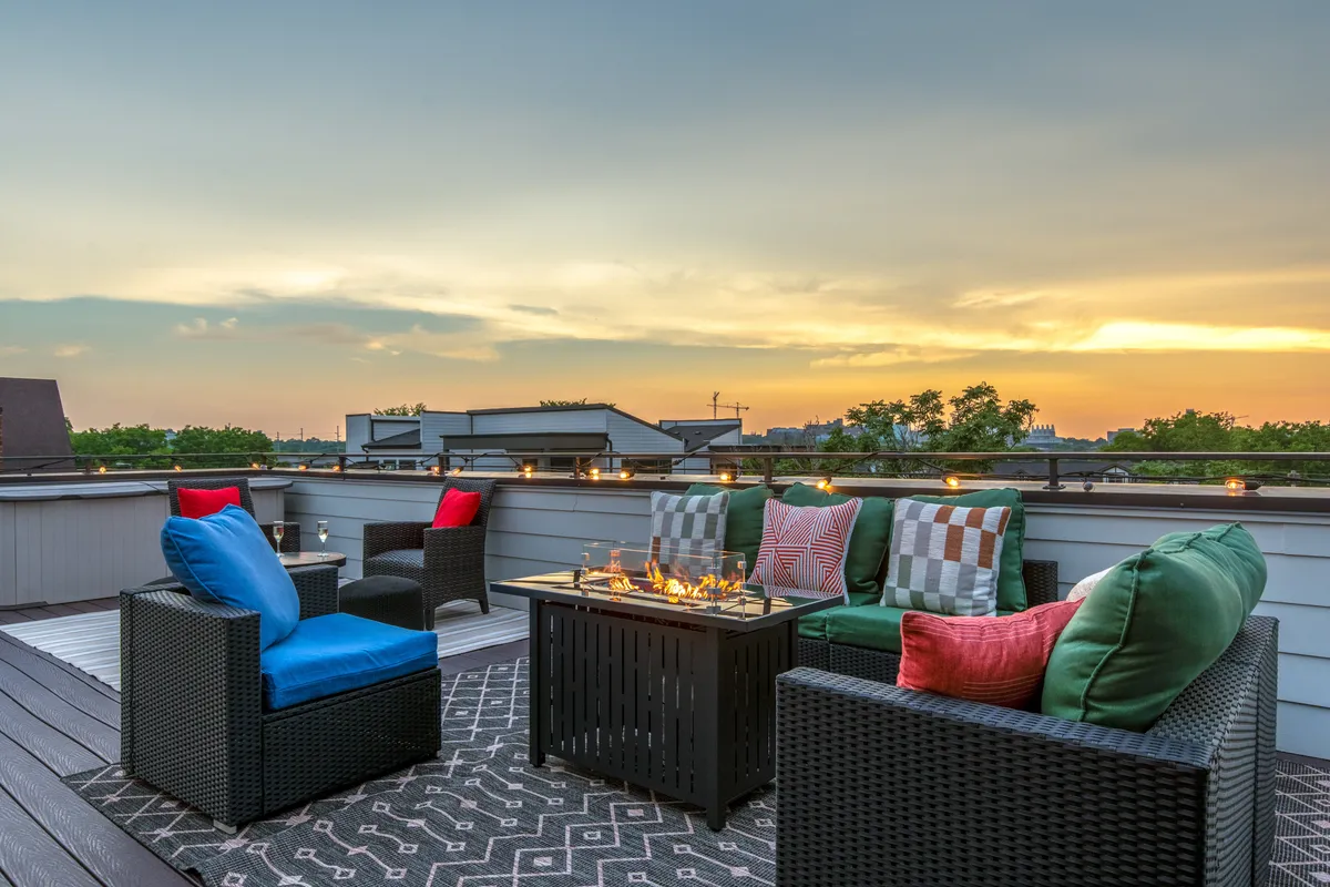 A cozy rooftop deck featuring a fire table surrounded by colorful cushions, with a sweeping view of the city skyline in the background. The scene is set for evening gatherings, illuminated by the warm glow from the fire and the ambient city lights.