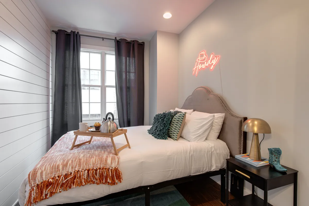 A welcoming bedroom with a bed dressed in decorative pillows, a neon sign, and a tray with teapot, setting a mood for relaxing mornings or peaceful reading sessions. The room feels thoughtfully designed for comfort and style.