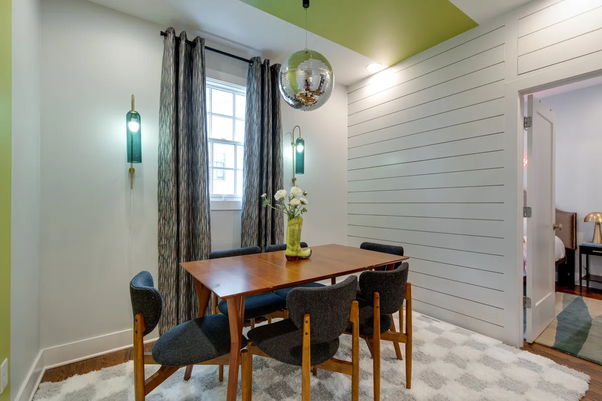 A stylish dining area anchored by a sleek table, statement hanging light fixture, disco ball, textured curtains, and a green accent wall. The setting is intimate yet lively, suited for memorable dinners, game nights, or celebratory meals.